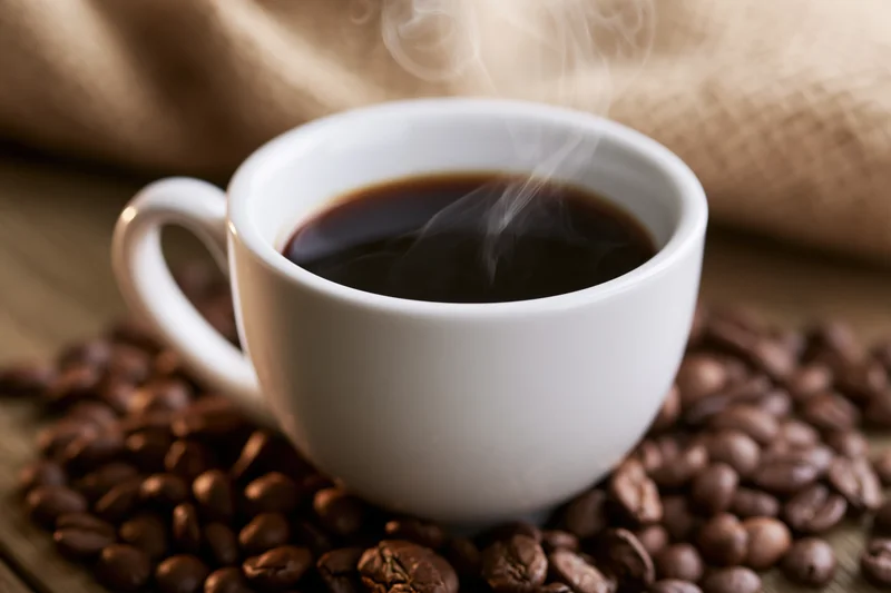 White ceramic cup filled with steaming dark coffee resting on roasted beans on wooden surface