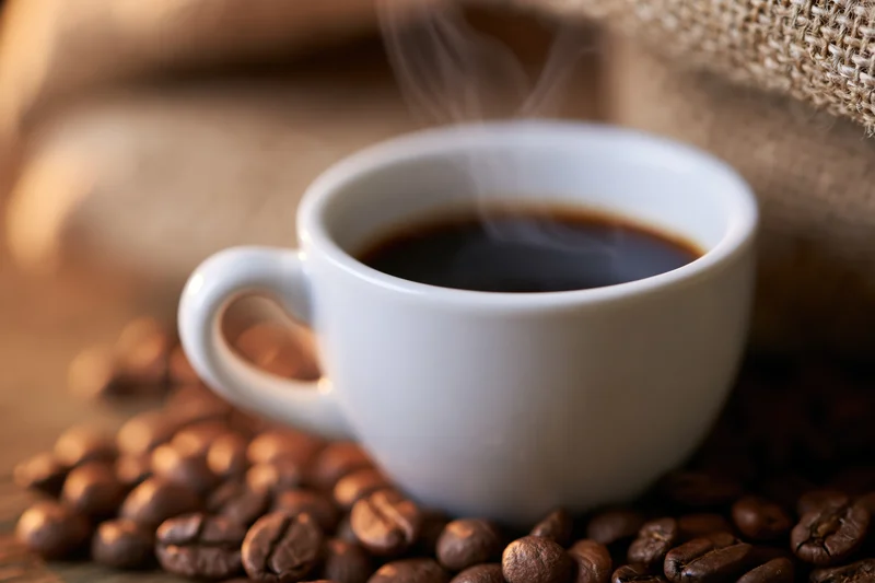 White ceramic espresso cup filled with steaming dark coffee resting on roasted beans on wooden surface