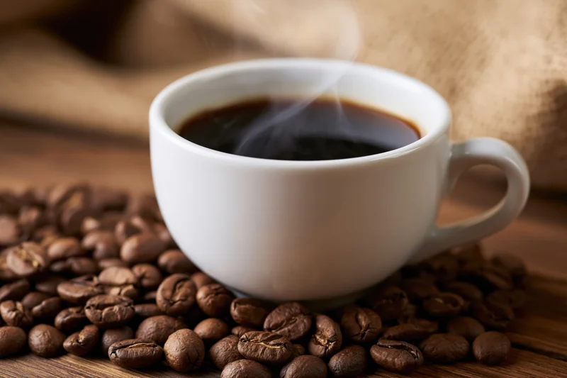 White ceramic cup filled with steaming dark coffee resting on roasted beans on wooden surface