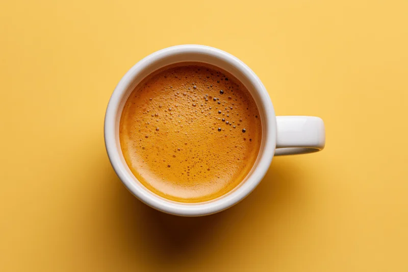 Overhead view of a white ceramic espresso cup filled with frothy coffee on a yellow background