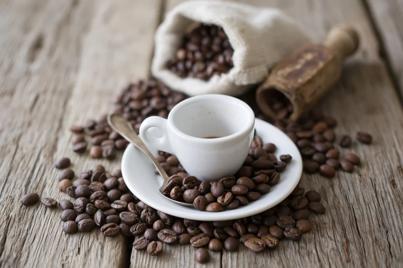White ceramic espresso cup on saucer with spoon surrounded by coffee beans on wooden table