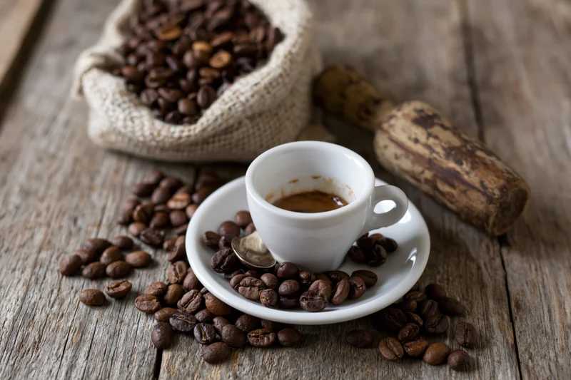 White ceramic espresso cup on saucer with scattered coffee beans and wooden grinder on rustic wooden table