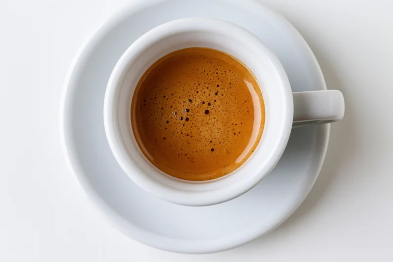 Overhead view of a white ceramic espresso cup with crema on a matching saucer on a white surface