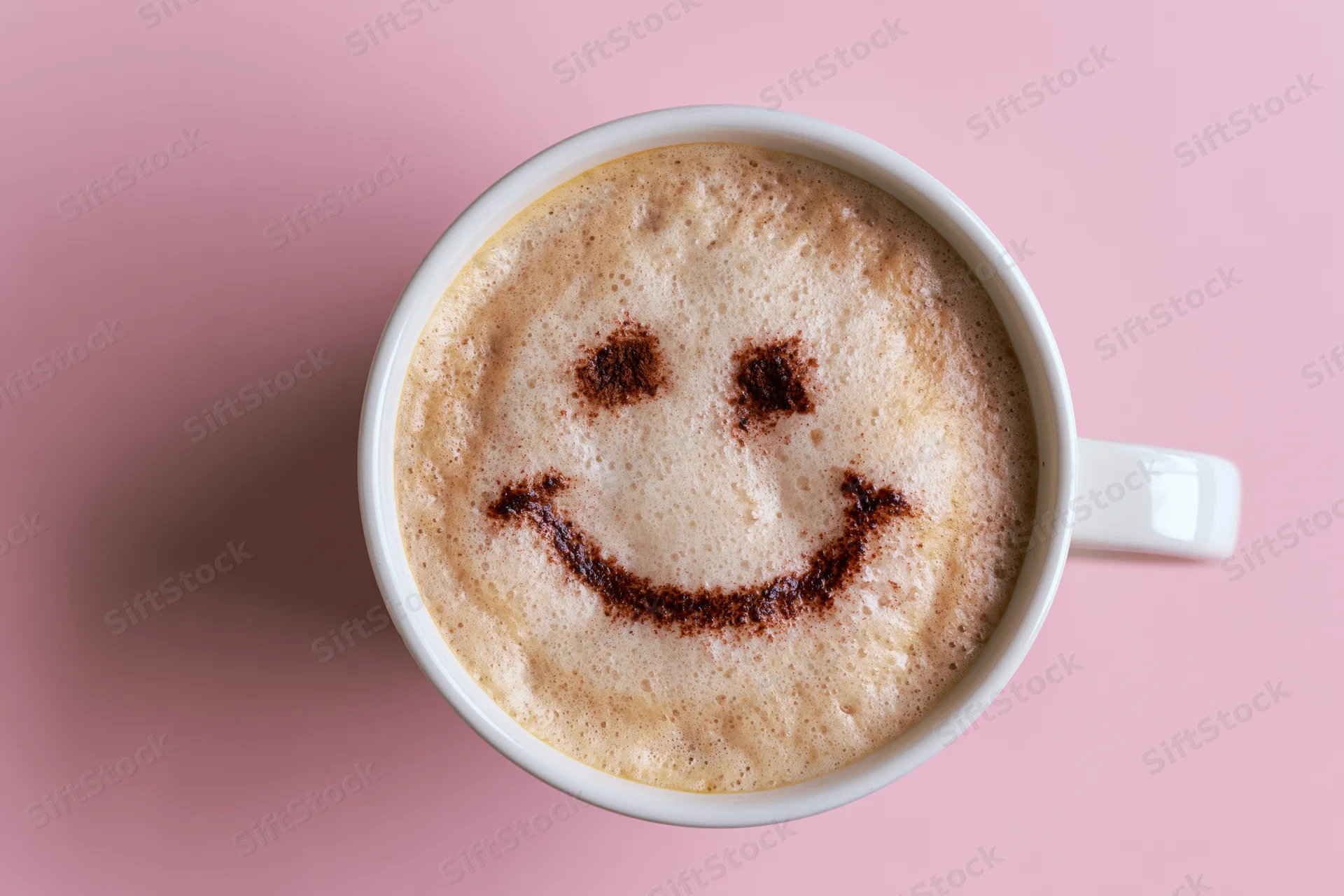 White ceramic mug filled with frothy cappuccino featuring a smiley face made of cinnamon on pink background