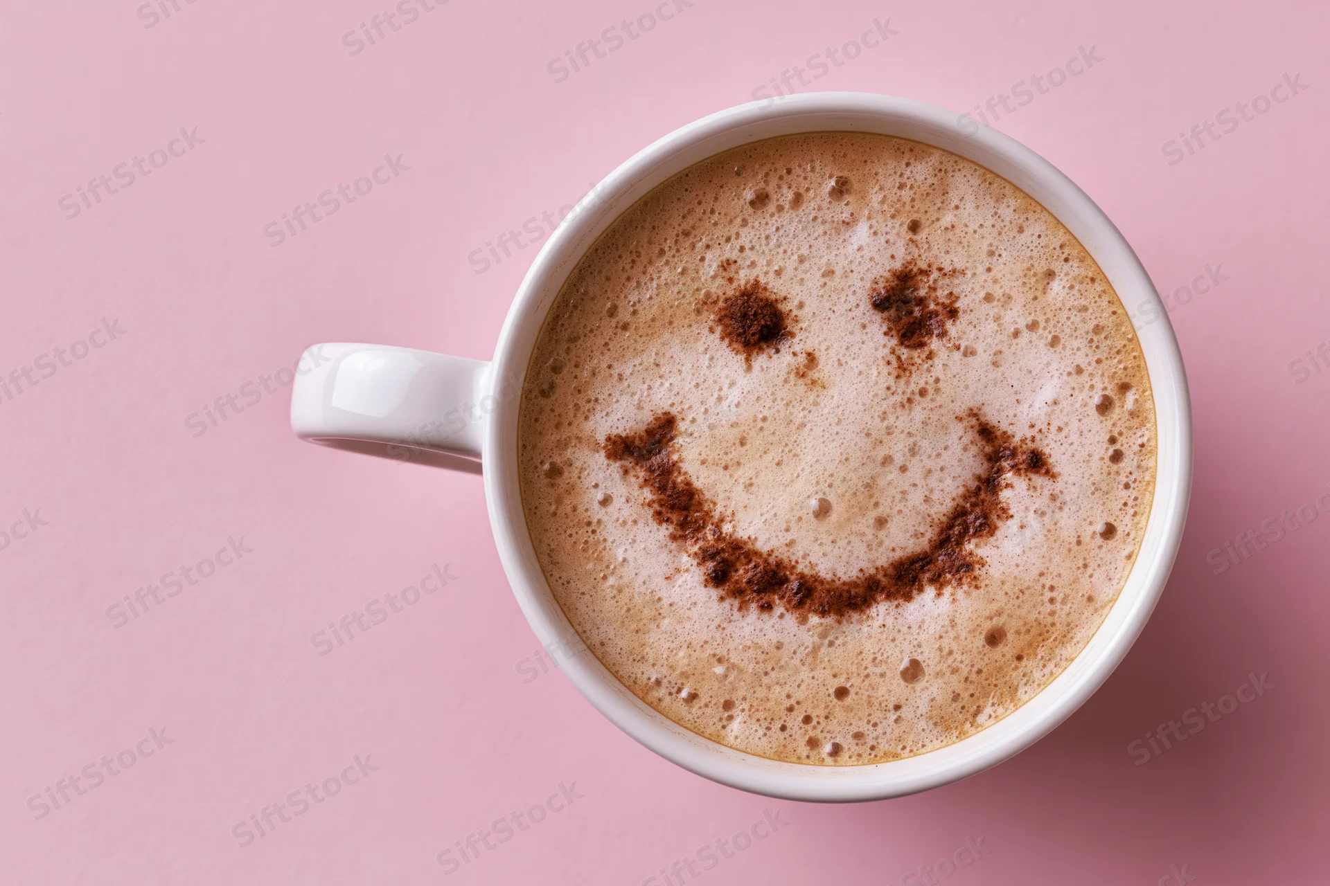 White ceramic mug filled with frothy cappuccino featuring a smiley face made of cinnamon on pink background