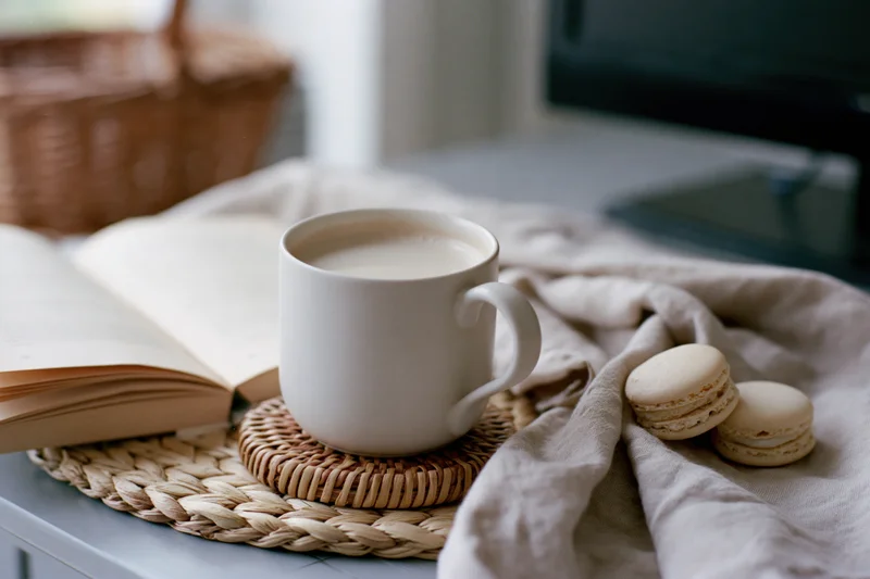 White ceramic mug of milky coffee on woven rattan trivet beside open book and macaroons on cloth-covered table