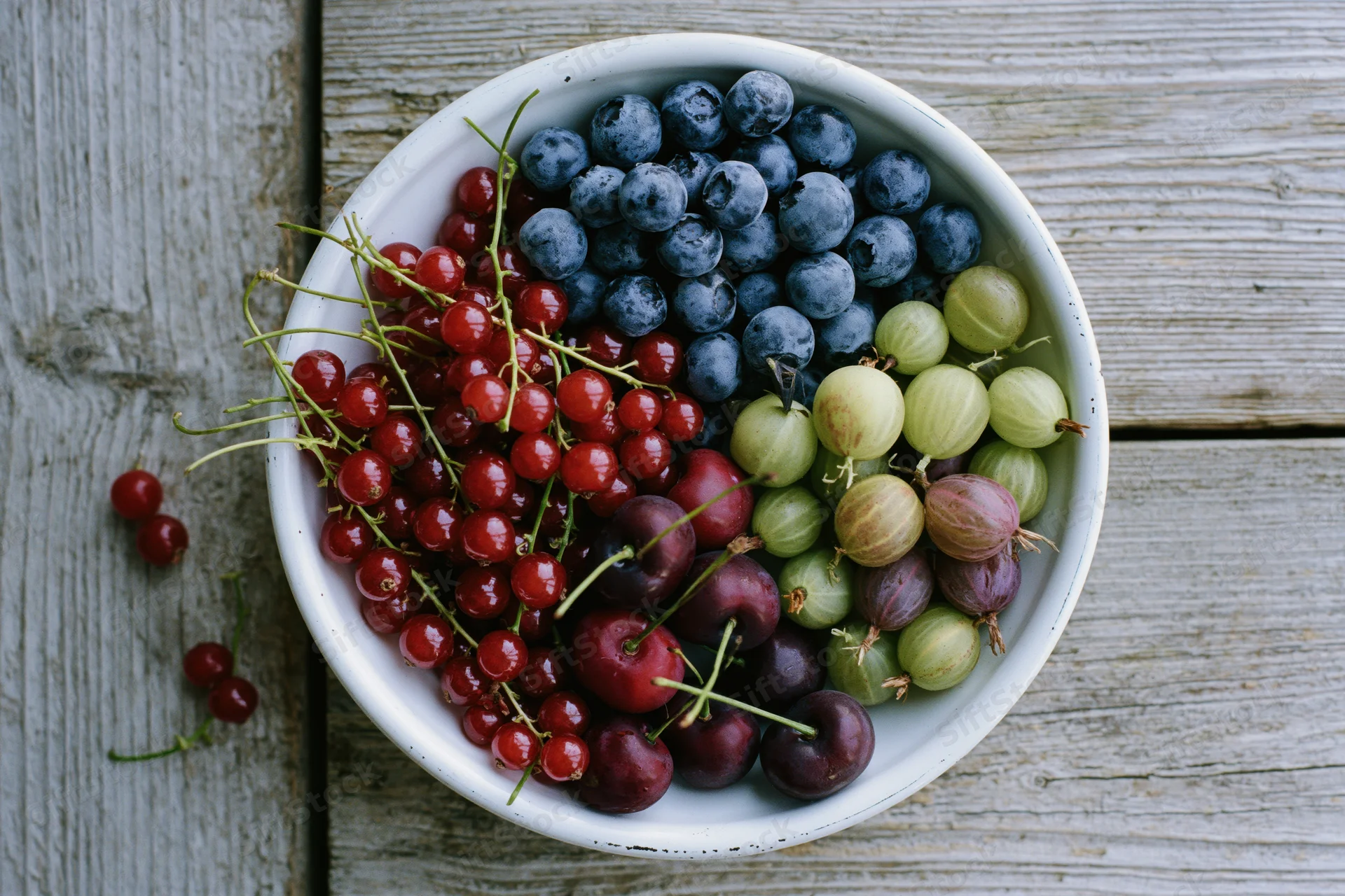 White enamel bowl filled with mixed summer berries on weathered wooden surface