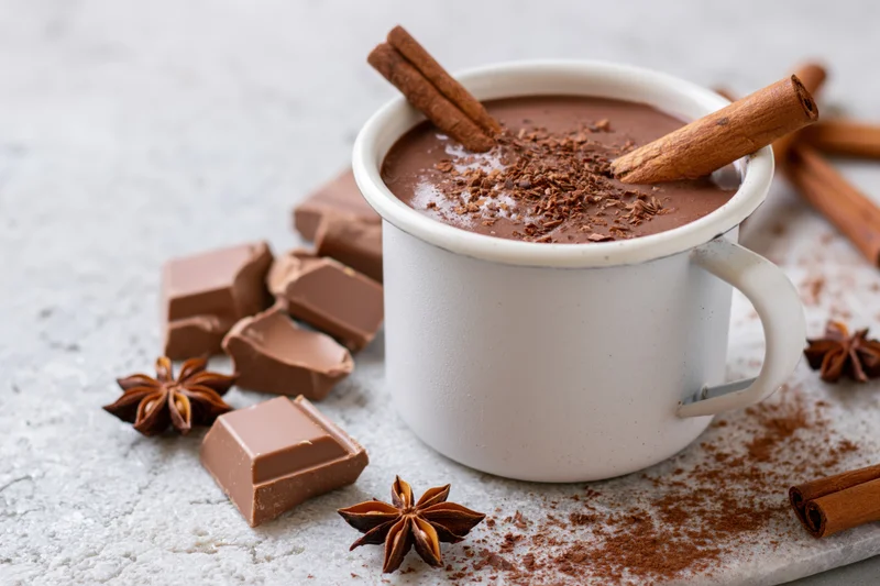 White enamel mug filled with thick hot chocolate topped with cinnamon sticks and chocolate shavings on a textured surface with scattered chocolate pieces and star anise