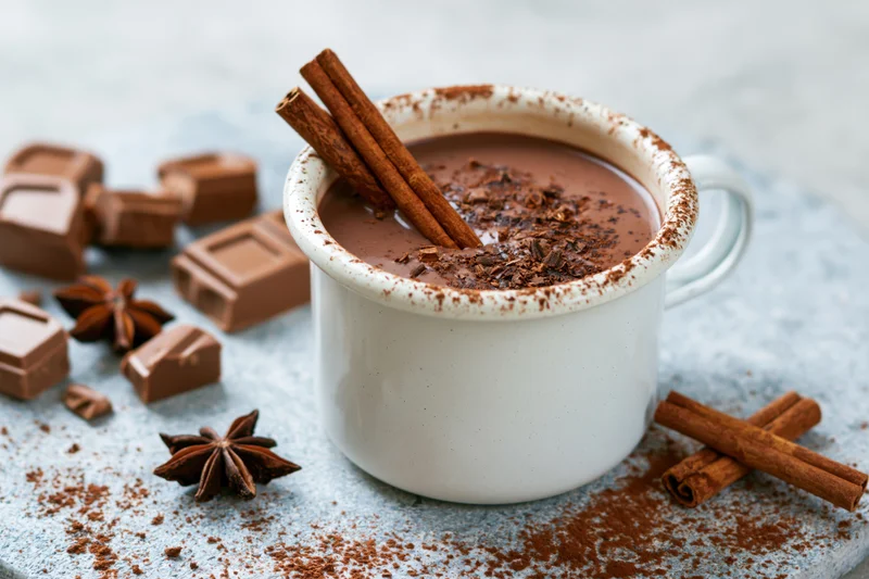 White enamel mug filled with hot chocolate topped with cinnamon sticks and cocoa powder on a textured surface with scattered chocolate pieces and star anise