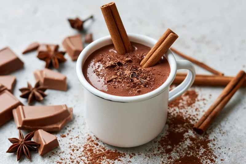 White enamel mug filled with thick hot chocolate topped with cinnamon sticks and chocolate shavings on a textured surface with scattered cocoa powder, chocolate chunks, and star anise around it