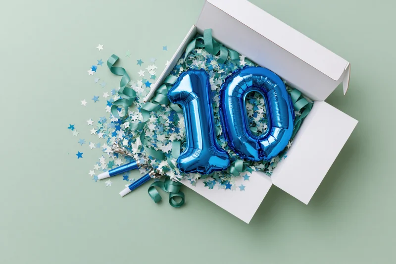 Blue number ten balloons in an open white gift box with confetti and party poppers on a mint green surface