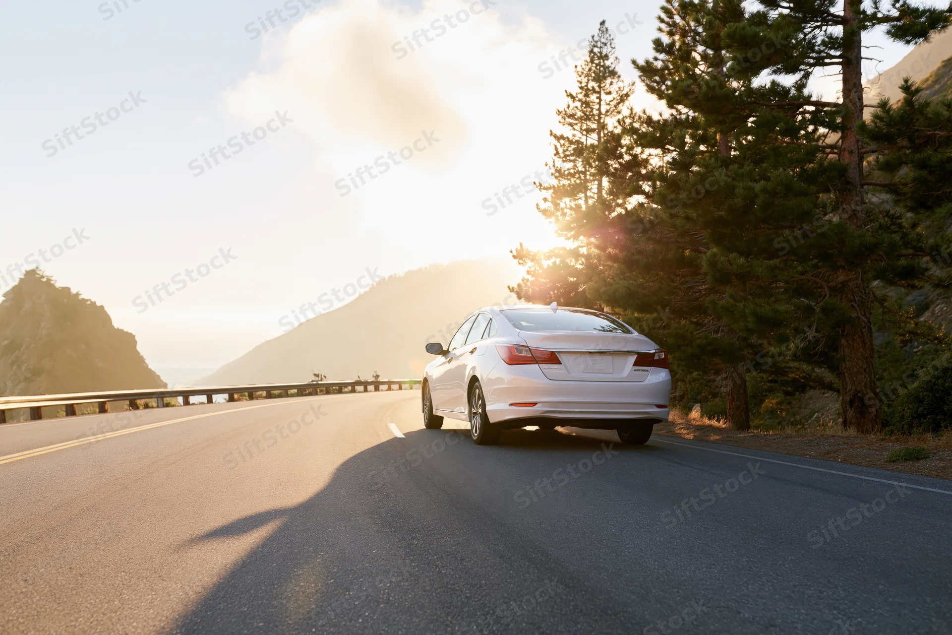 White sedan driving along winding coastal mountain highway at sunset