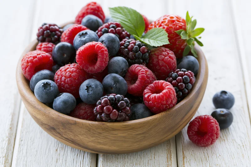 Mixed fresh berries in a wooden bowl on a white wooden surface