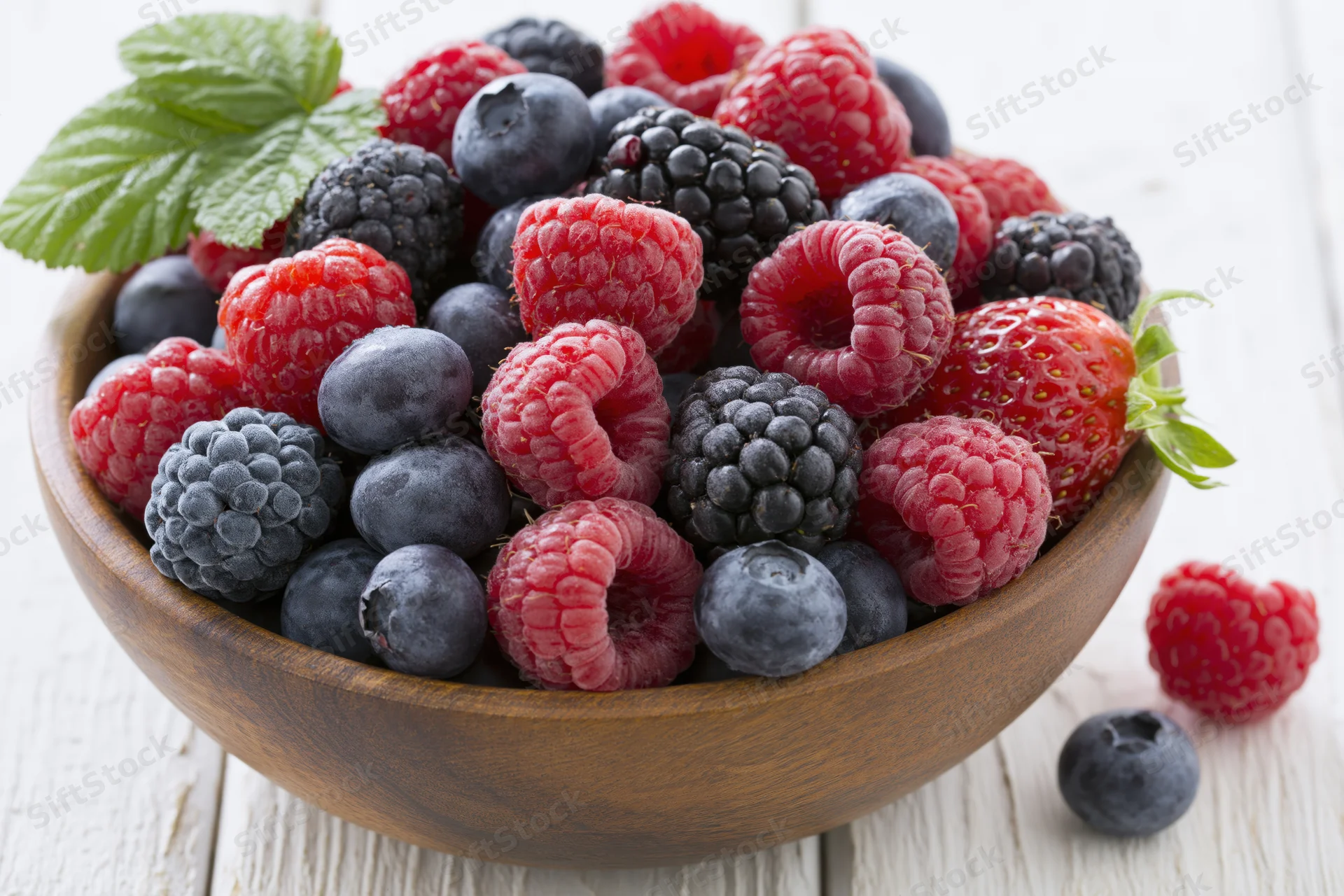 Mixed fresh berries in a wooden bowl on a white wooden surface