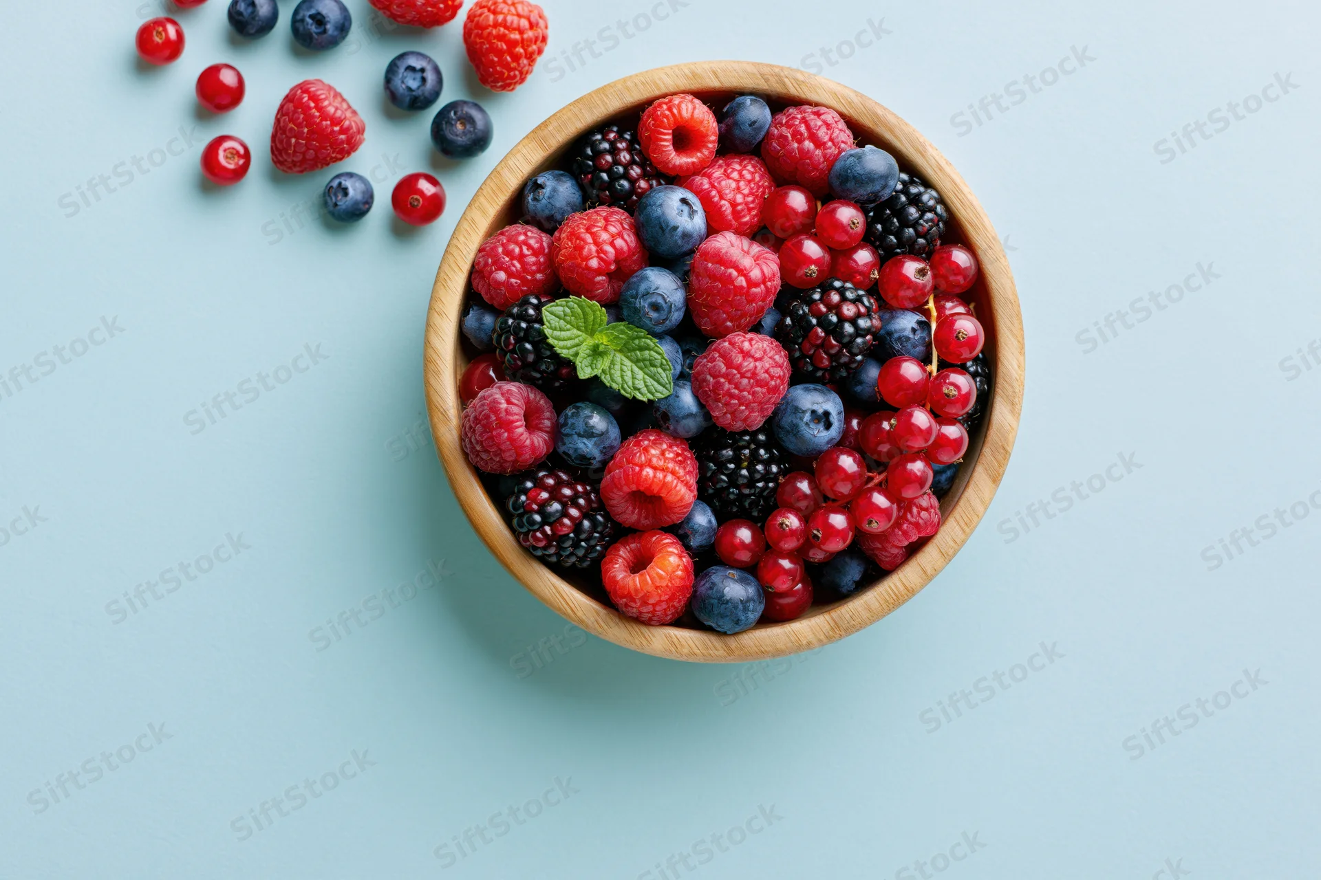 Wooden bowl filled with mixed fresh berries and mint on light blue surface