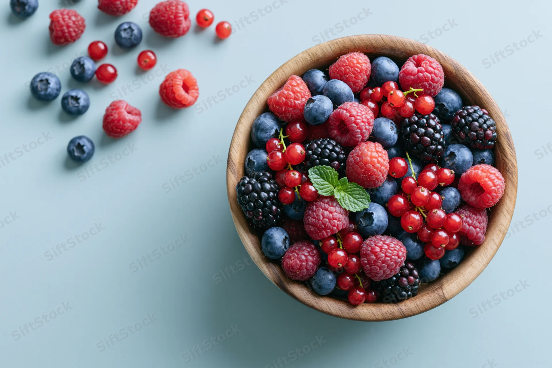 Mixed berries in a wooden bowl on a light blue surface with scattered fruit and mint garnish