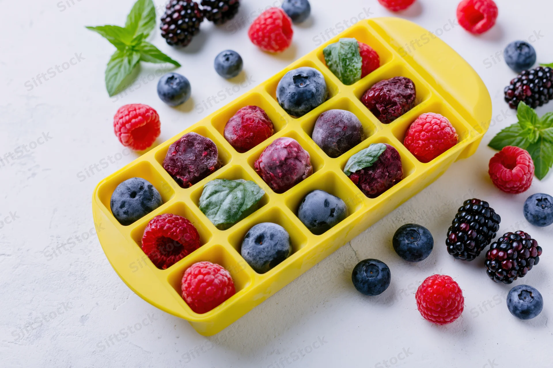 Frozen mixed berries in a yellow plastic ice cube tray on a white surface with scattered fruit and mint leaves