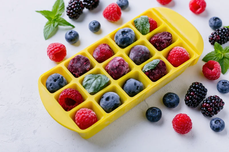 Frozen mixed berries in a yellow plastic ice cube tray on a white surface with scattered fruit and mint leaves