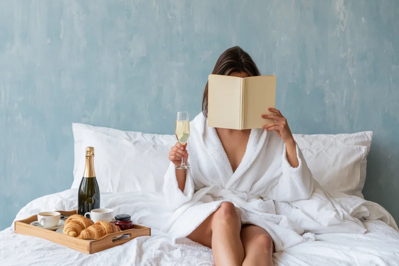 Woman in white bathrobe reading book in bed with breakfast tray and champagne