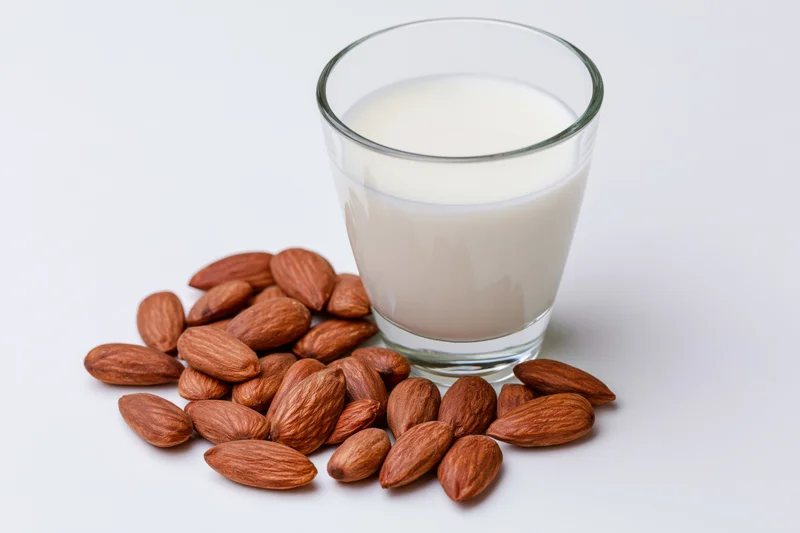 Glass of almond milk beside scattered almonds on white surface