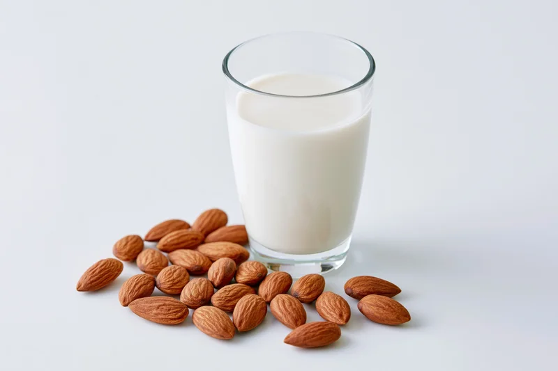 Glass of almond milk beside scattered almonds on white surface