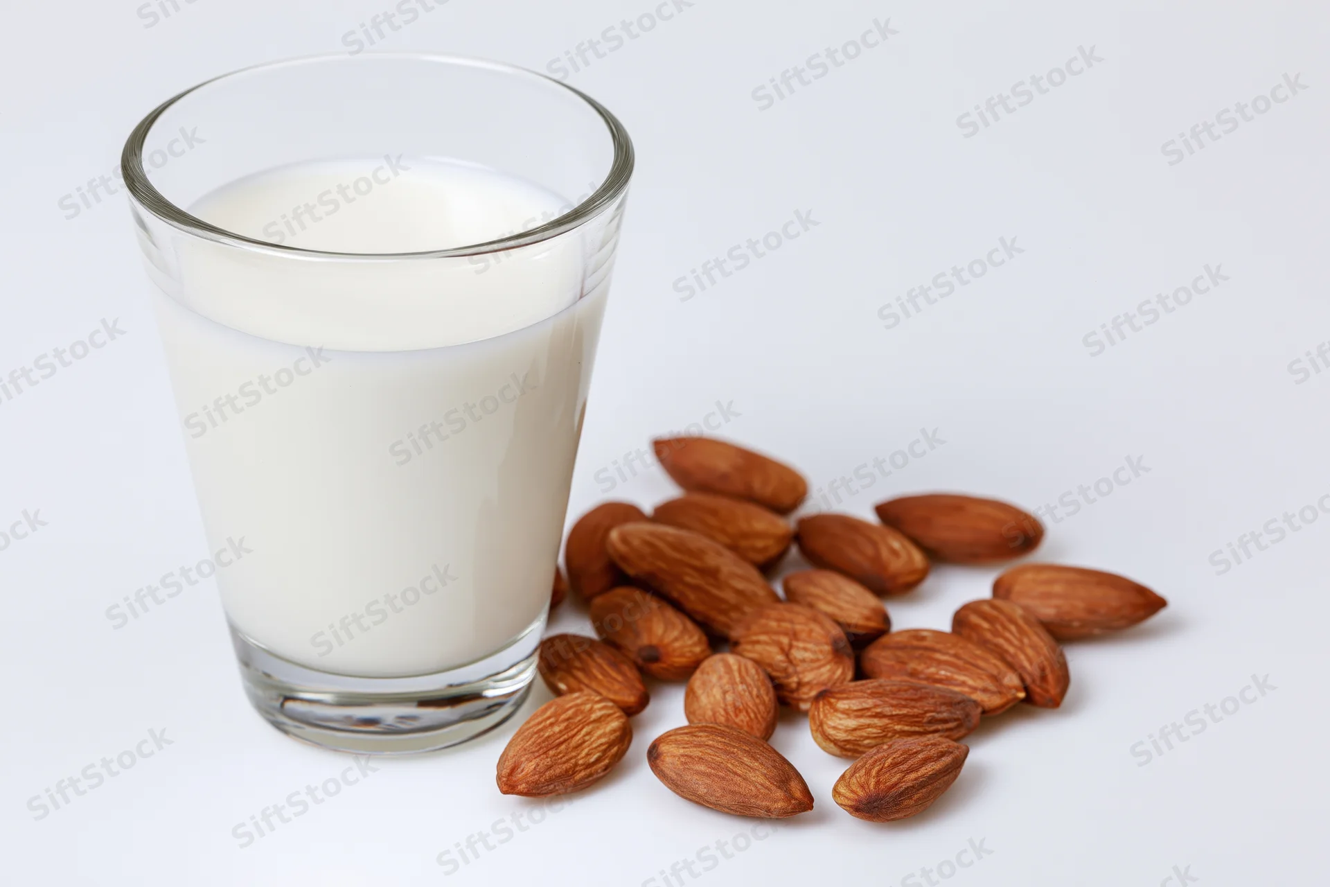 Glass of almond milk beside scattered almonds on white surface