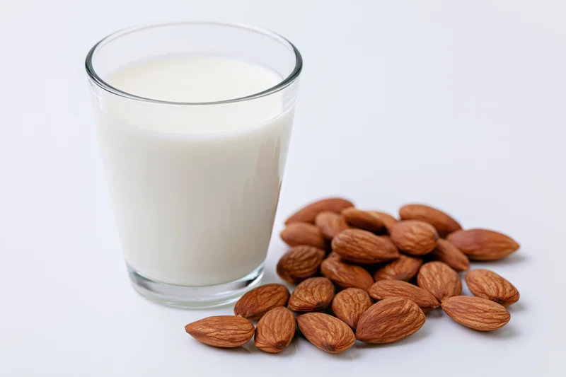 Glass of almond milk beside scattered almonds on white surface