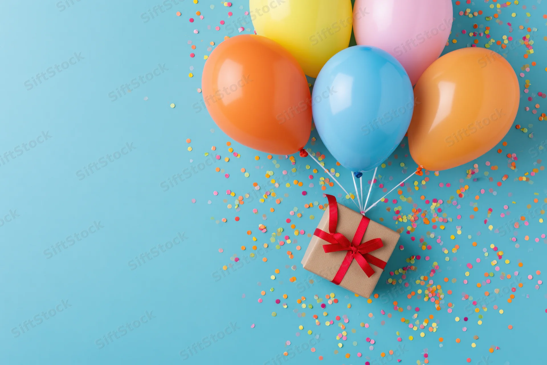 Colorful balloons tied to a gift box with red ribbon on a blue surface scattered with confetti