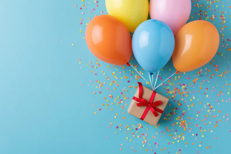 Colorful balloons tied to a gift box with red ribbon on a blue surface scattered with confetti