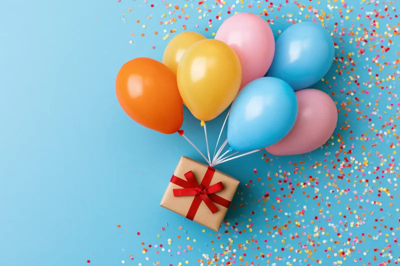 Colorful balloons tied to a gift box with red ribbon on blue background scattered with confetti