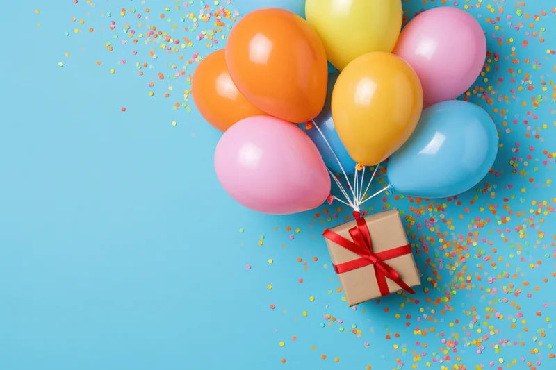 Colorful balloons tied to a gift box with red ribbon on a blue background scattered with confetti
