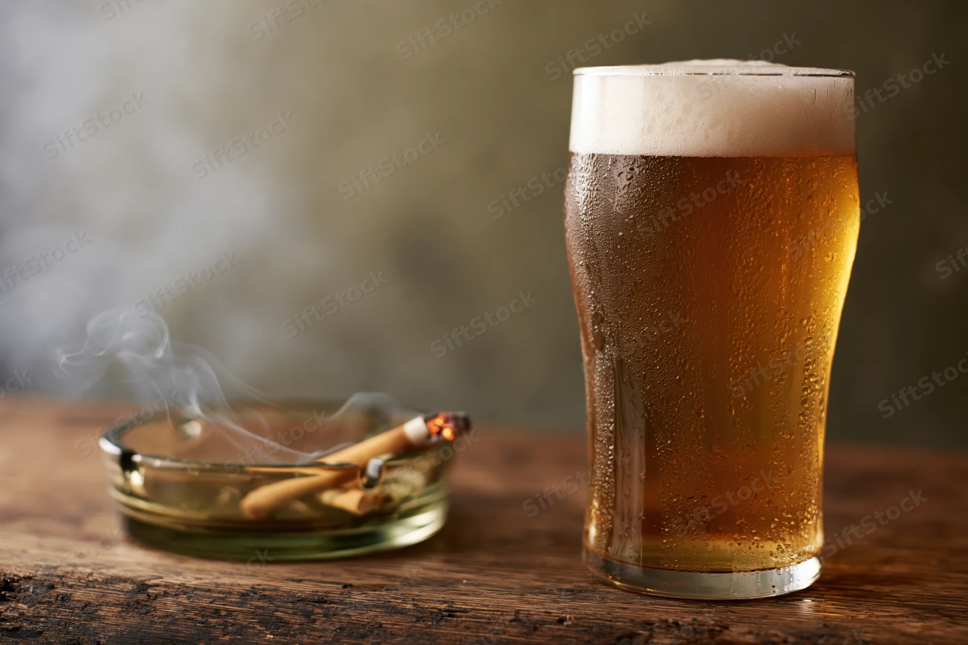 Cold glass of pale lager with foamy head and condensation beside a smoking cigarette in an ashtray on a wooden table
