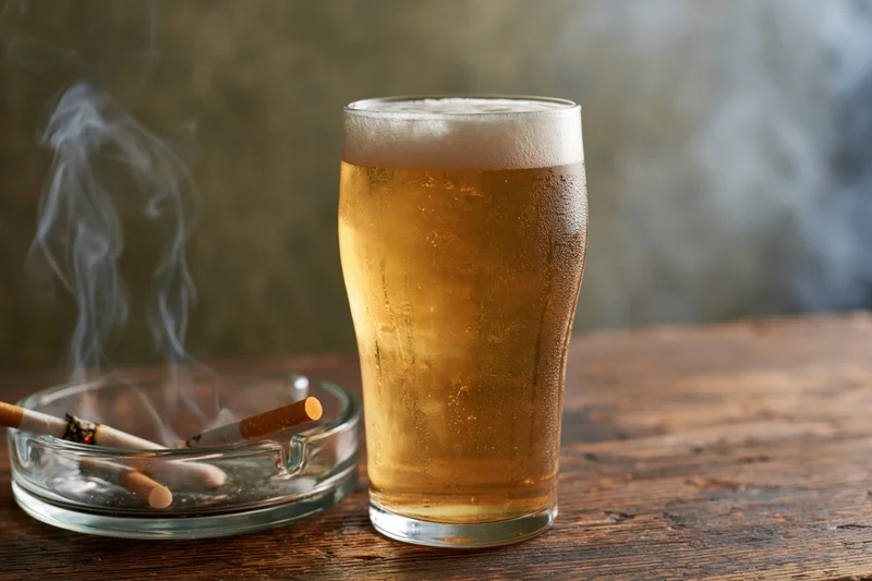 Cold pint of pale lager with foamy head and smoking cigarette in ashtray on wooden table