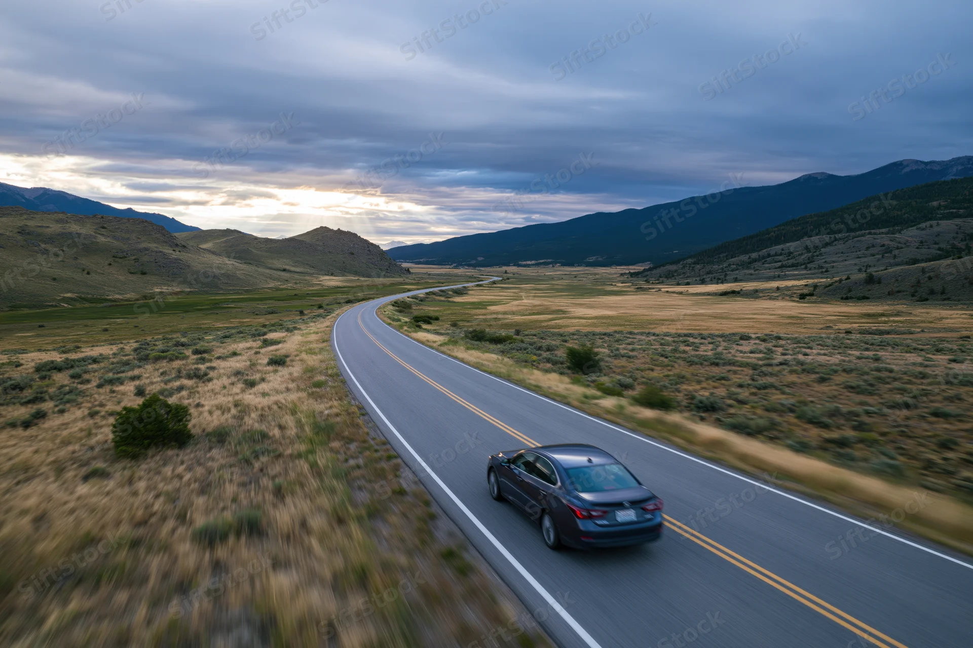 Dark sedan driving away on winding two-lane highway through rural landscape with mountains and cloudy sky at dusk
