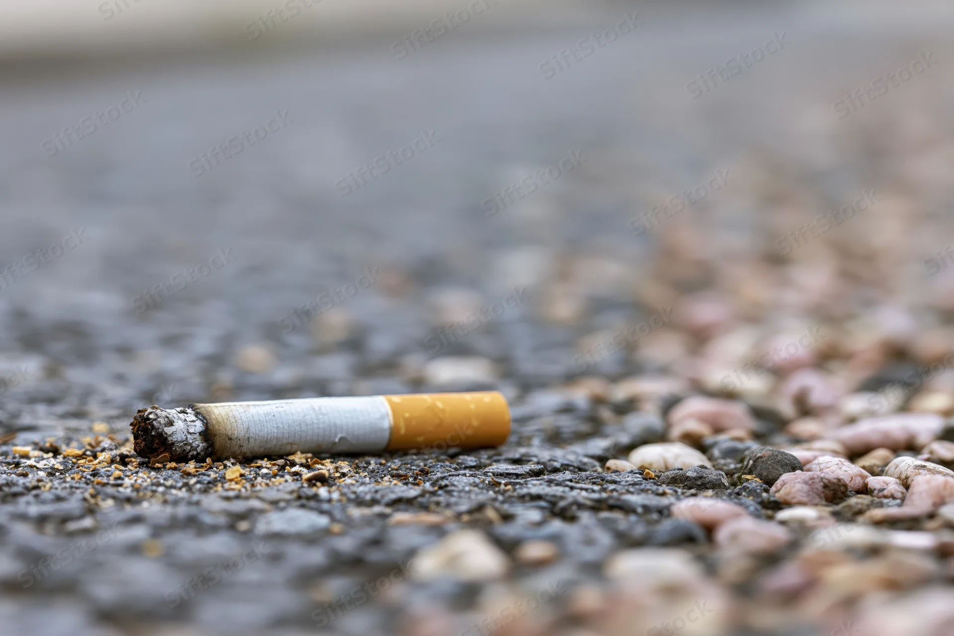 Discarded cigarette butt on rough asphalt pavement with gravel and ash