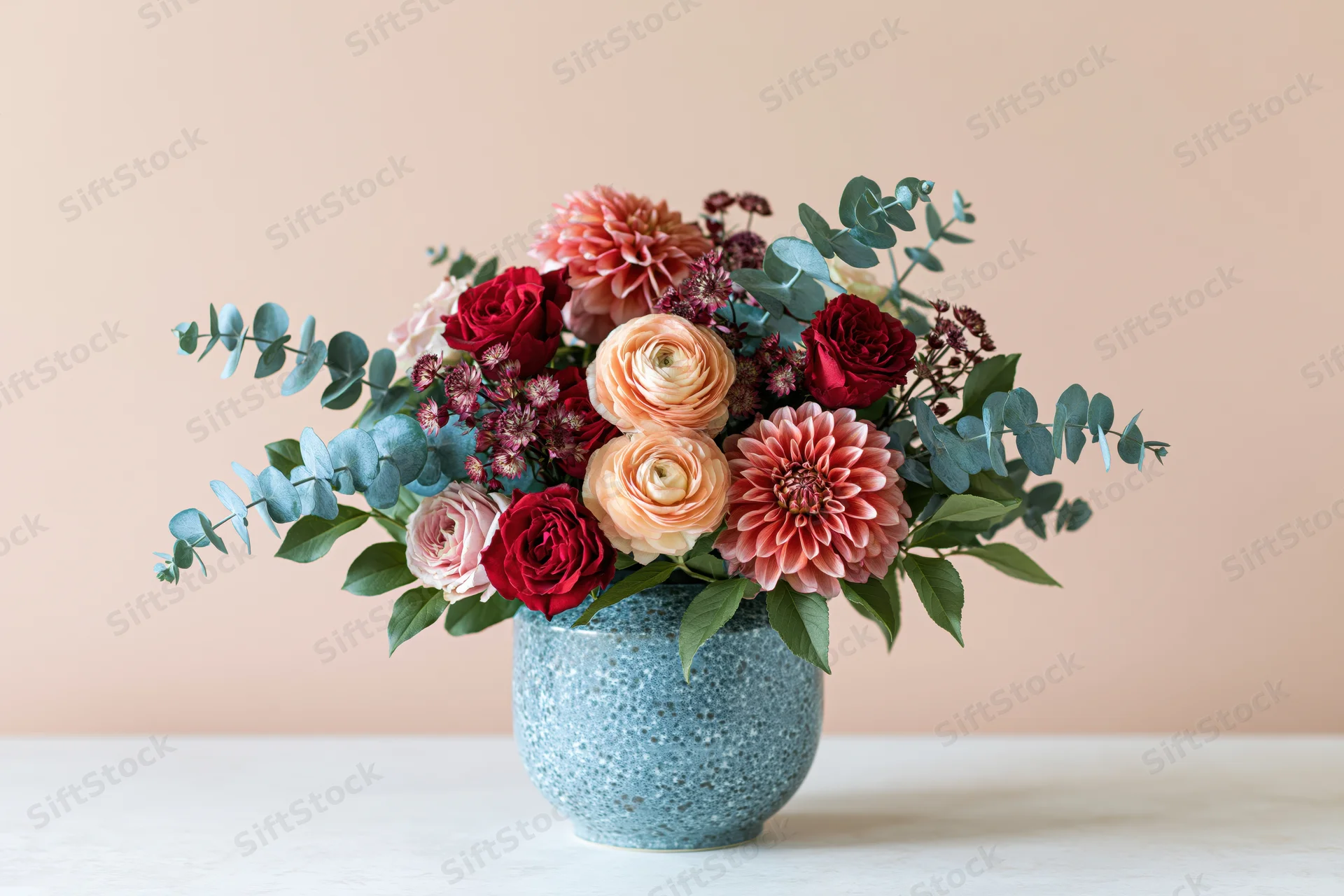 Floral bouquet with ranunculus, roses, dahlias and eucalyptus in a blue speckled ceramic vase on a white table