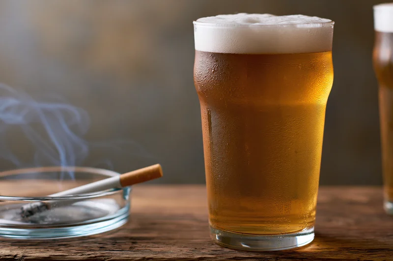 Full pint of golden lager with thick foam head on wooden table beside smoking cigarette in ashtray