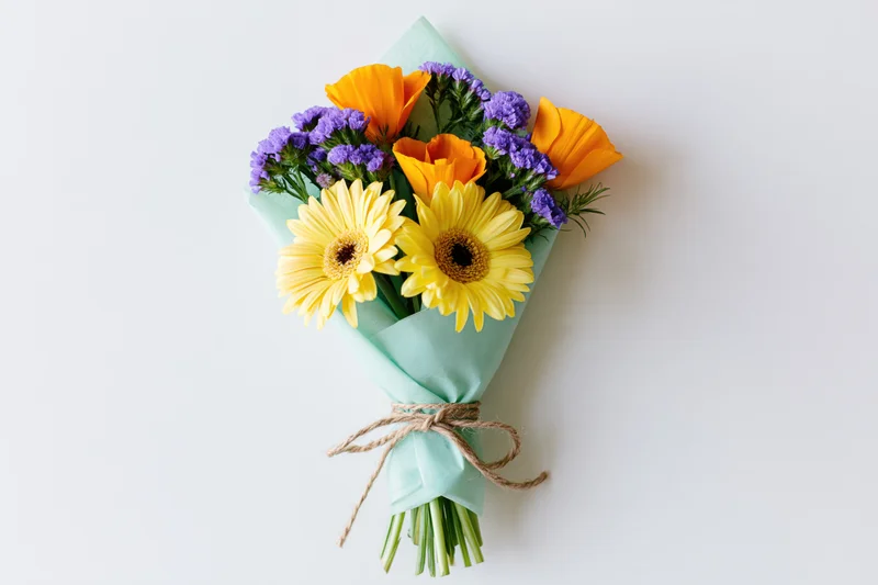 Hand-tied bouquet of yellow gerbera daisies, orange poppies, and purple statice wrapped in mint paper with twine on white surface