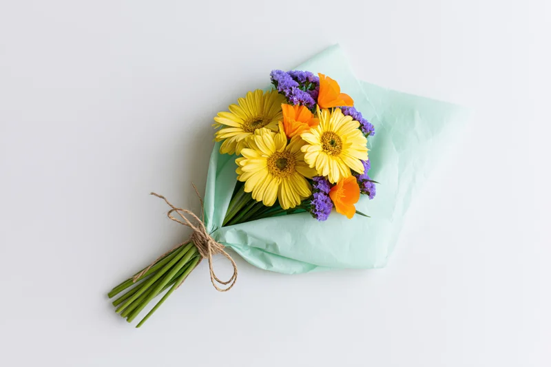 Hand-tied bouquet of yellow gerbera daisies, orange and purple flowers wrapped in mint paper on white surface