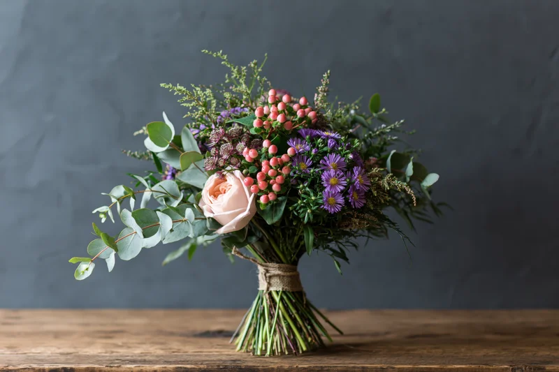 Hand-tied wildflower bouquet with eucalyptus and pink berries on a wooden table against a dark gray background