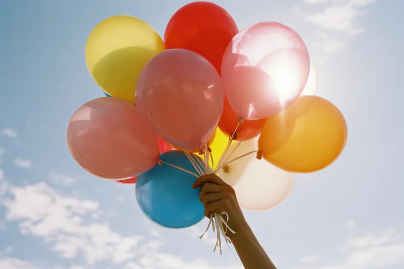 Hand holding a bunch of colorful latex balloons against a blue sky with clouds