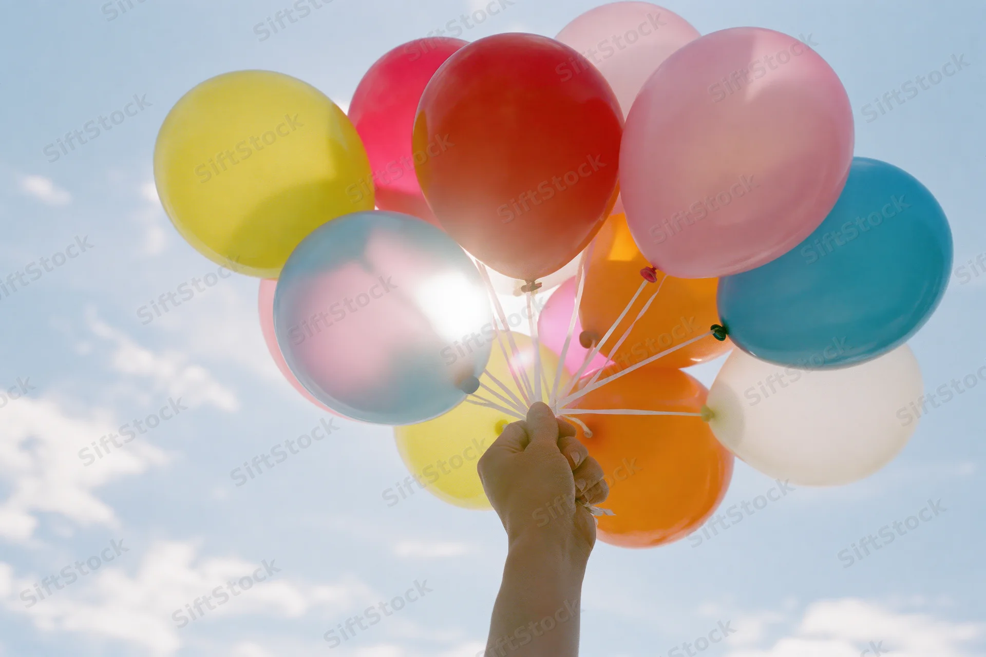 Hand gripping a large bunch of colorful latex balloons against a blue sky with clouds