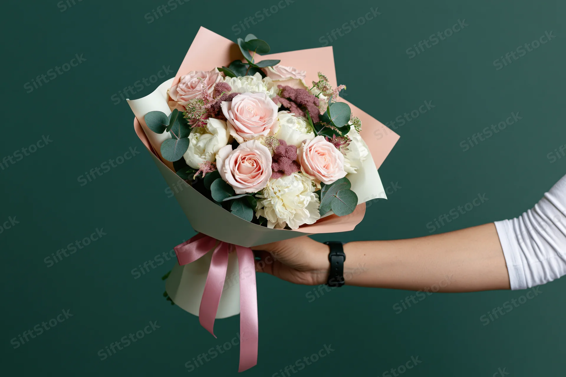 Woman's hand holding a wrapped bouquet of pink roses and white peonies with eucalyptus against a dark green background