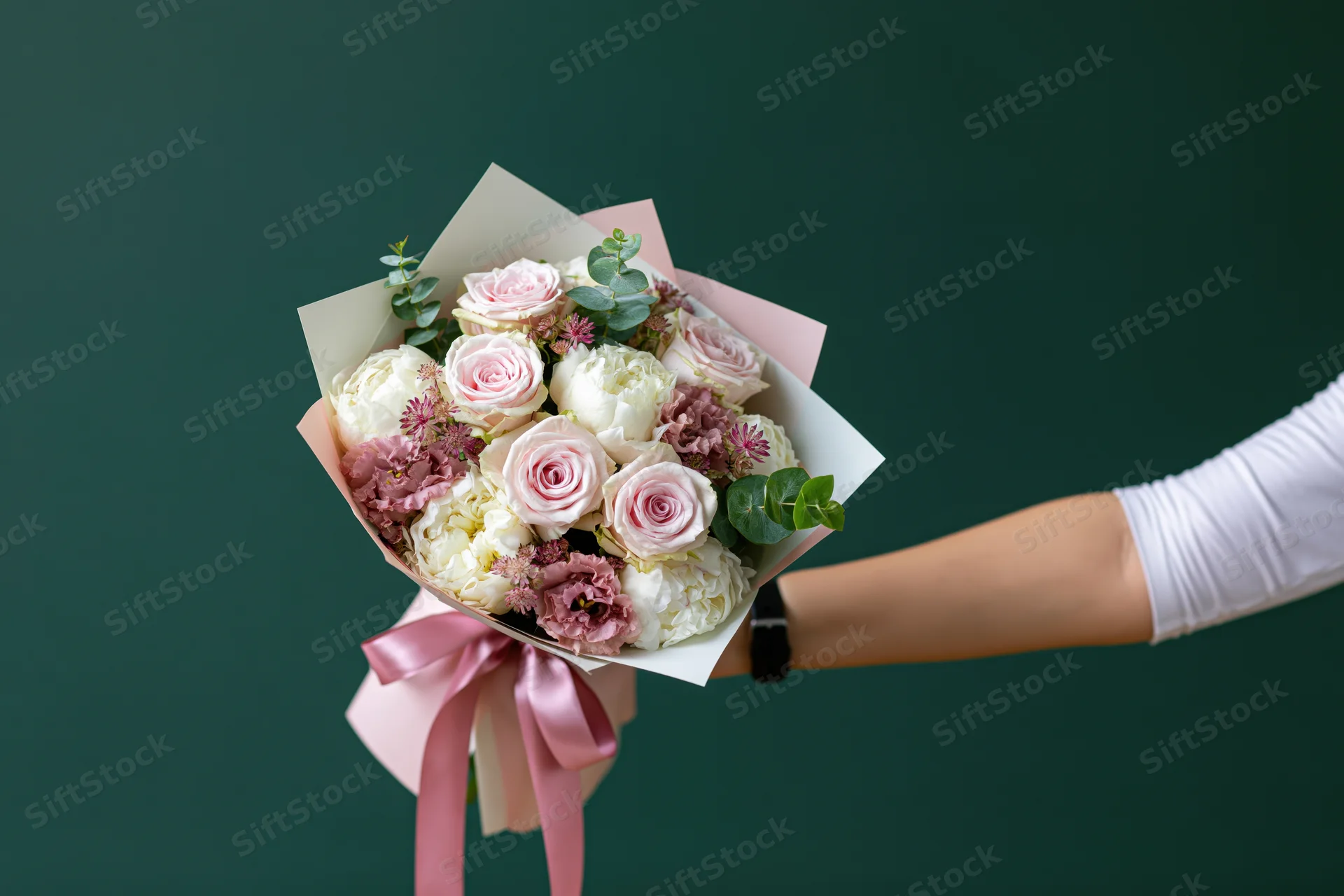 Woman's hand holding a wrapped bouquet of pink roses and white peonies against a green background