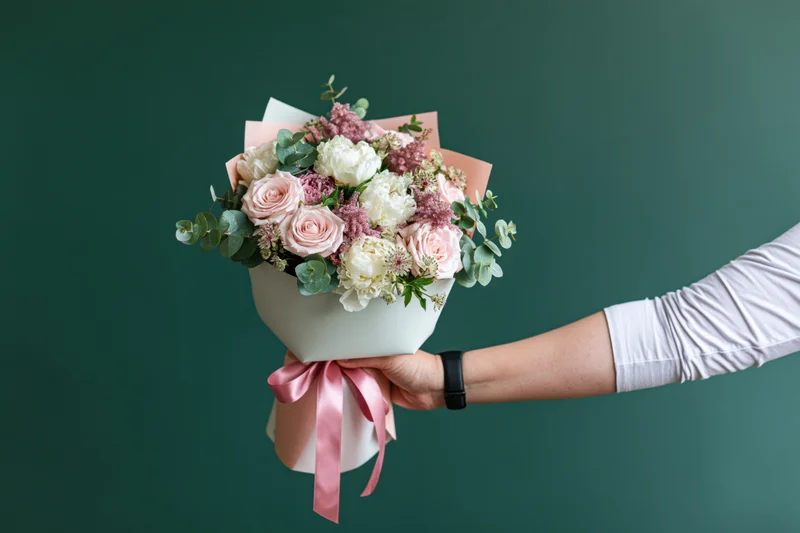 Woman's hand holding a wrapped bouquet of pink and white roses and peonies with eucalyptus against a green background