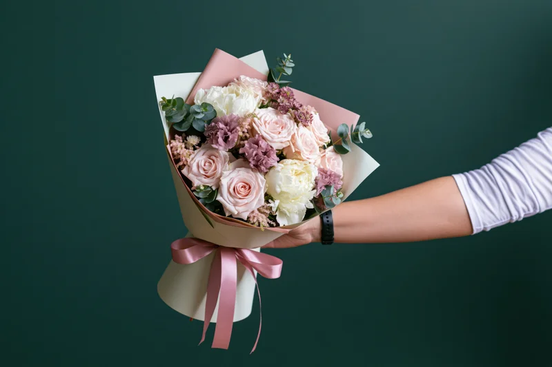 Hand holding a wrapped bouquet of pink and white roses with eucalyptus against a green background
