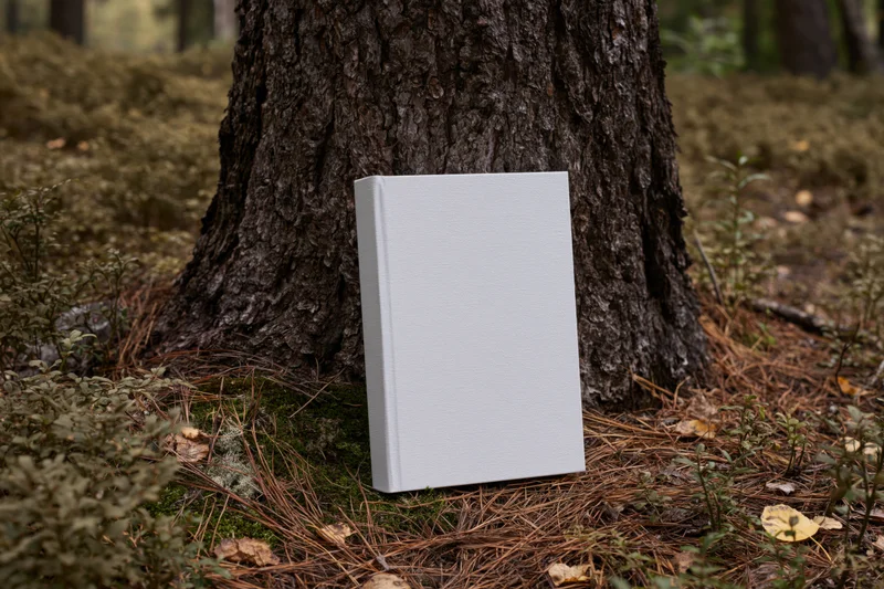 Hardcover book with plain white blank cover leaning against tree trunk in forest setting