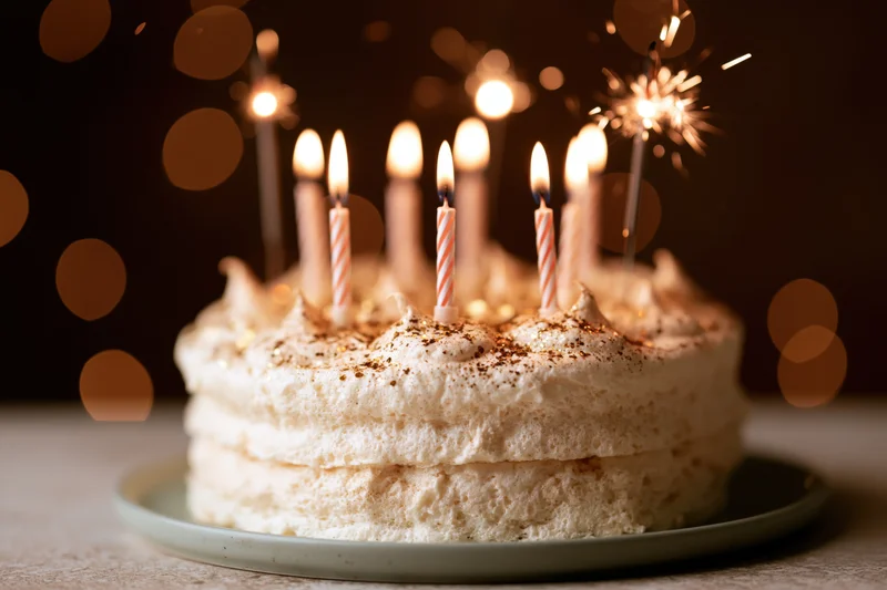 Layered cream birthday cake with lit striped candles and sparkler on plate