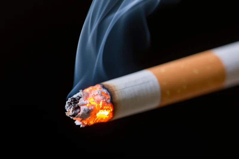 Lit cigarette burning with glowing ember and rising smoke against black background