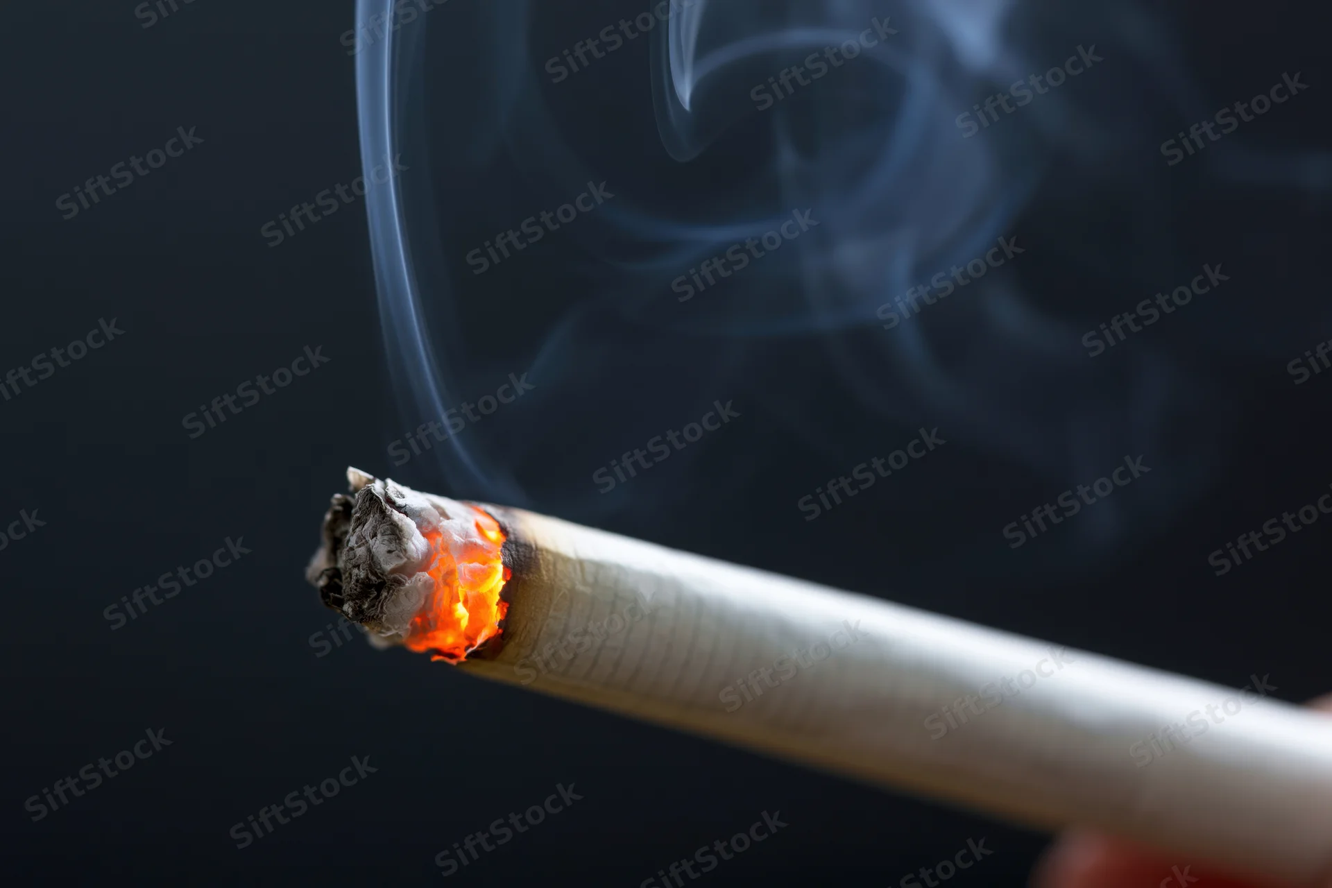 Lit cigarette with glowing ember and curling smoke held in hand against dark background